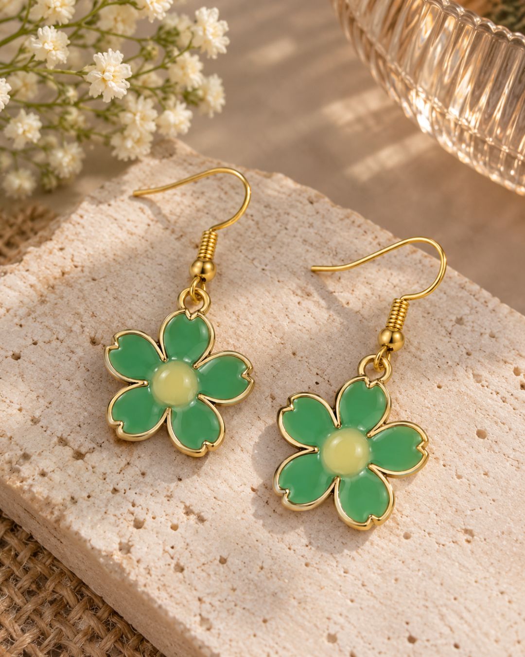 A pair of gold-tone flower dangle earrings with green enamel petals and pale yellow centers, displayed on a textured stone slab with small white flowers and a glass bowl in the background.