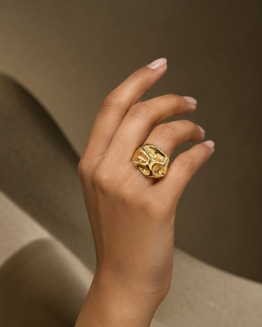 A close-up of a hand wearing a chunky, sculptural gold ring with organic, flowing textures, set against a warm, neutral, abstract background.