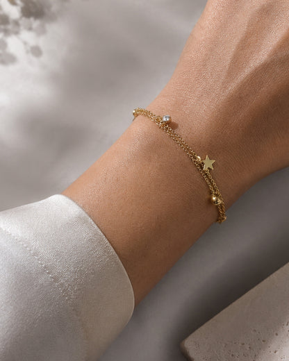 A delicate gold chain bracelet with a star charm and a small clear stone, shown worn on a person's wrist.