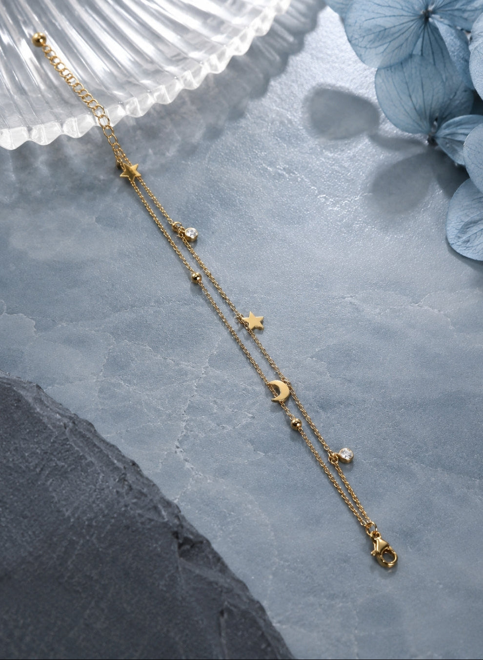 A delicate gold-toned double-strand chain necklace with celestial charms, including stars, moons, and small beads, styled on a textured gray surface with a white shell and blue flowers.