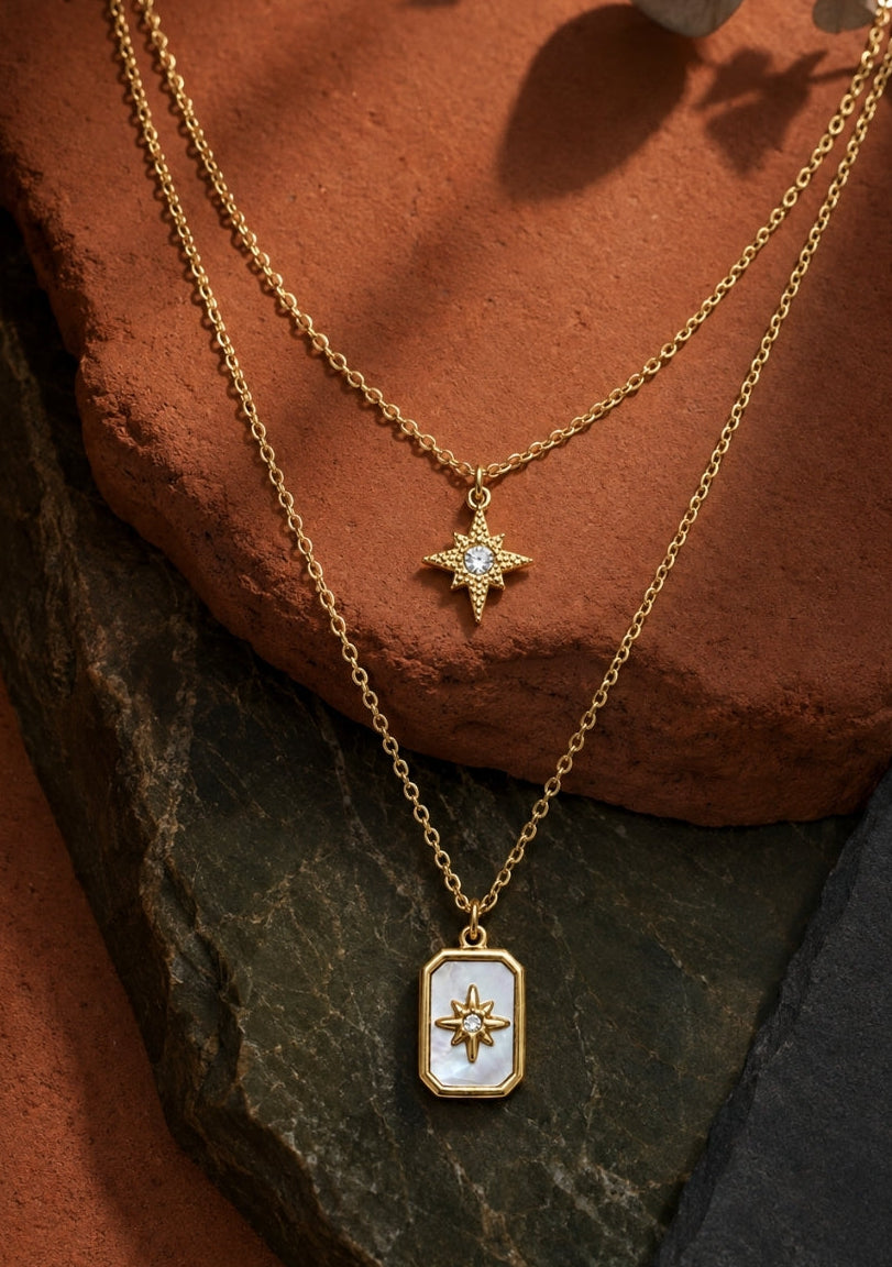 Two layered gold necklaces displayed on textured terracotta and dark stone. The shorter chain features a small gold cross pendant with a clear stone, while the longer chain holds a rectangular gold pendant with a white inlay and a gold sunburst motif.