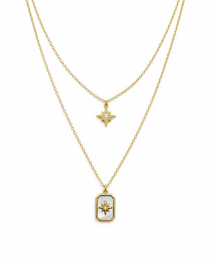 A double-layered gold necklace set on a white background. The shorter chain features a small gold starburst pendant with a clear stone, while the longer chain features a gold rectangular pendant with a white inlay and a gold sunburst design in the center.