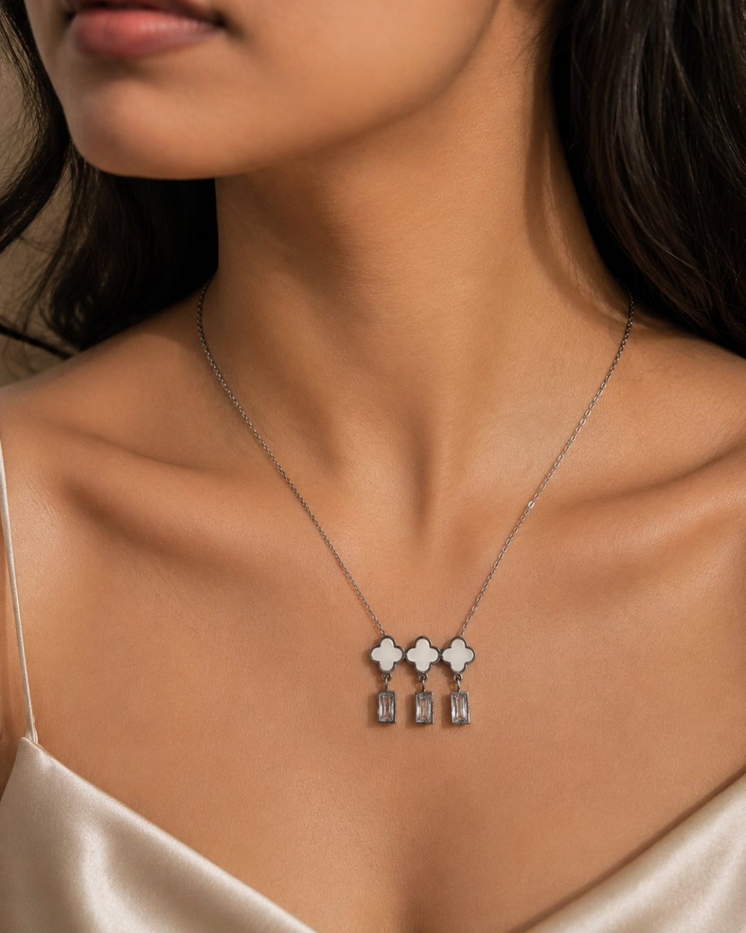 A close-up of a model wearing a delicate silver necklace with three clover-shaped pendants featuring white inlay and dangling clear crystals.