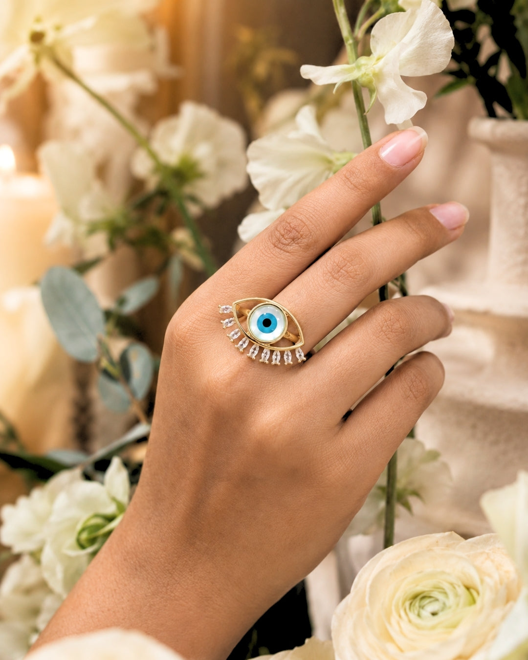 A gold ring featuring an evil eye motif with a blue center and small clear stones, worn on a hand with white flowers in the background.