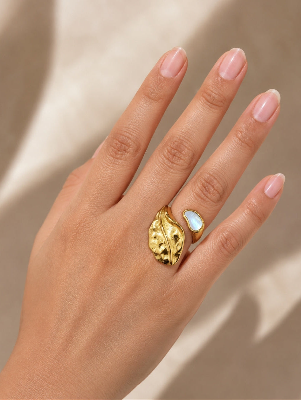 A close-up of a hand wearing a gold leaf-shaped ring with a white opal inlay, set against a warm beige background with soft shadows.