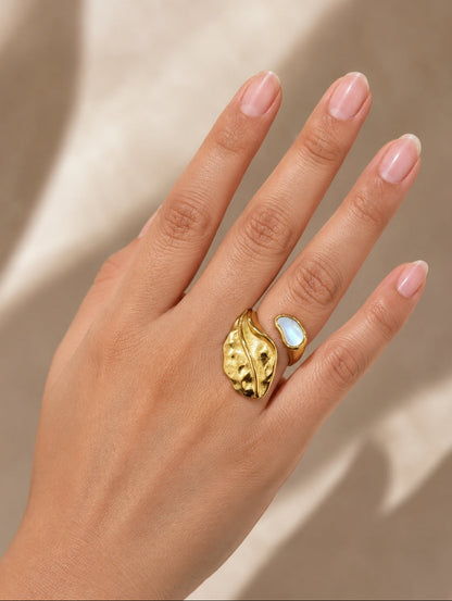 A close-up of a hand wearing a gold leaf-shaped ring with a white opal inlay, set against a warm beige background with soft shadows.