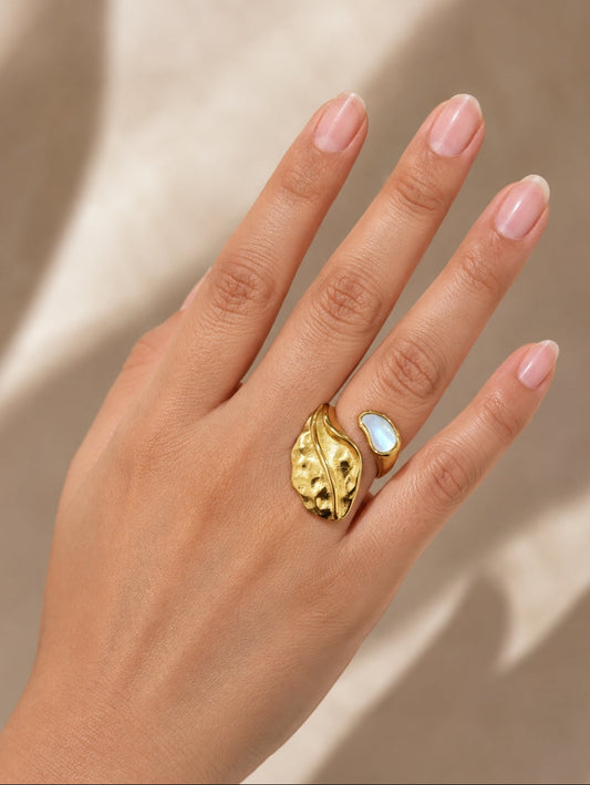 A close-up of a hand wearing a gold leaf-shaped ring with a white opal inlay, set against a warm beige background with soft shadows.