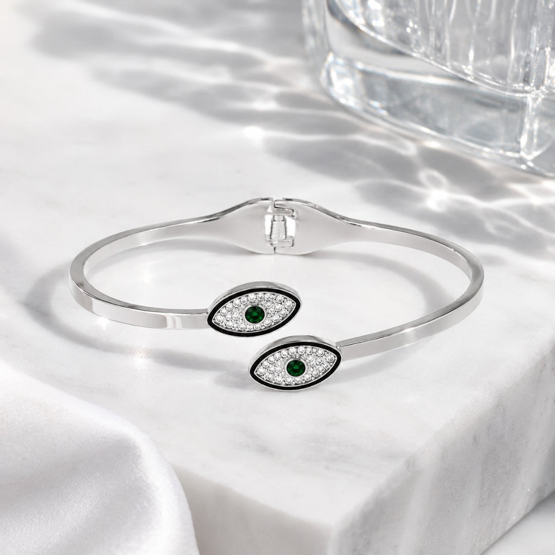 A silver cuff bracelet with two evil eye charms featuring green emerald centers and clear pavé accents, displayed on a white marble surface with soft shadows.