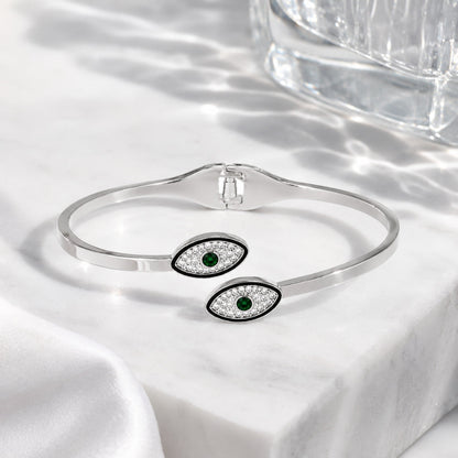 A silver cuff bracelet with two evil eye charms featuring green emerald centers and clear pavé accents, displayed on a white marble surface with soft shadows.