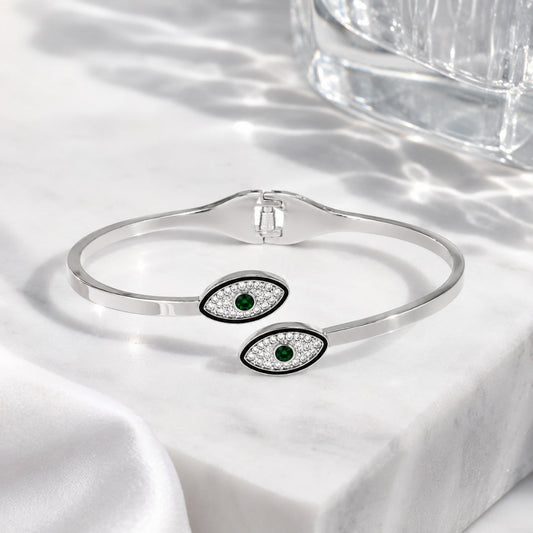 A silver cuff bracelet with two evil eye charms featuring green emerald centers and clear pavé accents, displayed on a white marble surface with soft shadows.