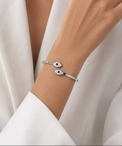 A close-up of a person's wrist wearing a slim silver cuff bracelet with two evil eye charms, styled with a white draped garment.