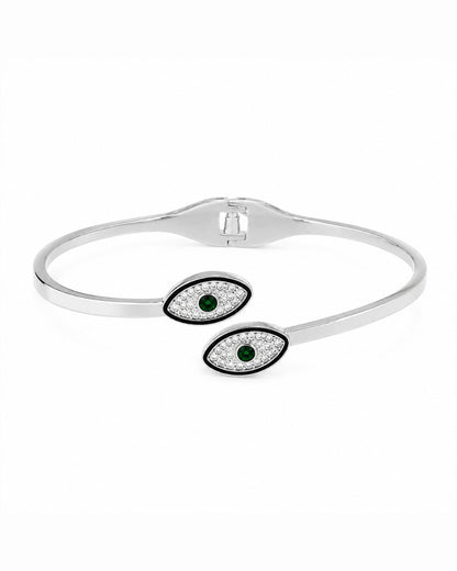 A silver-toned open cuff bangle bracelet featuring two evil eye charms with black borders, clear pavé stones, and green gemstone centers, shown on a plain white background.