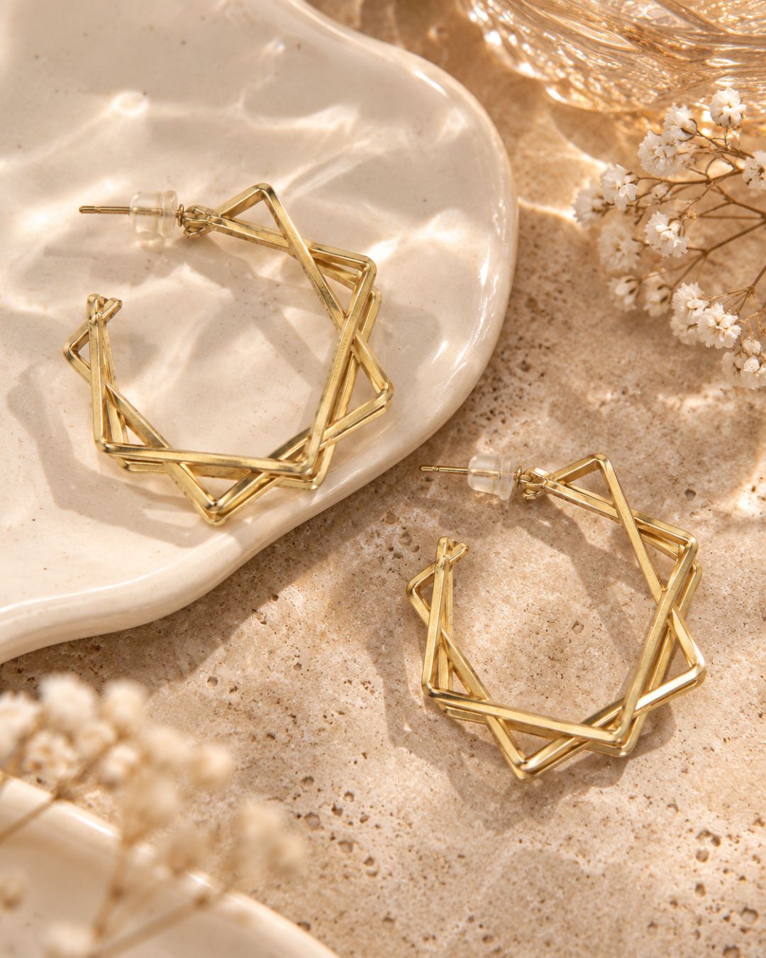 Gold geometric hoop earrings with an interlocking star design, displayed on a textured beige surface next to a white ceramic dish and dried white flowers.