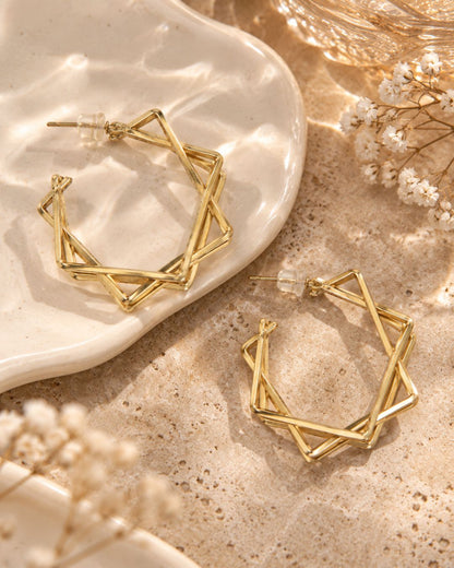 Gold geometric hoop earrings with an interlocking star design, displayed on a textured beige surface next to a white ceramic dish and dried white flowers.