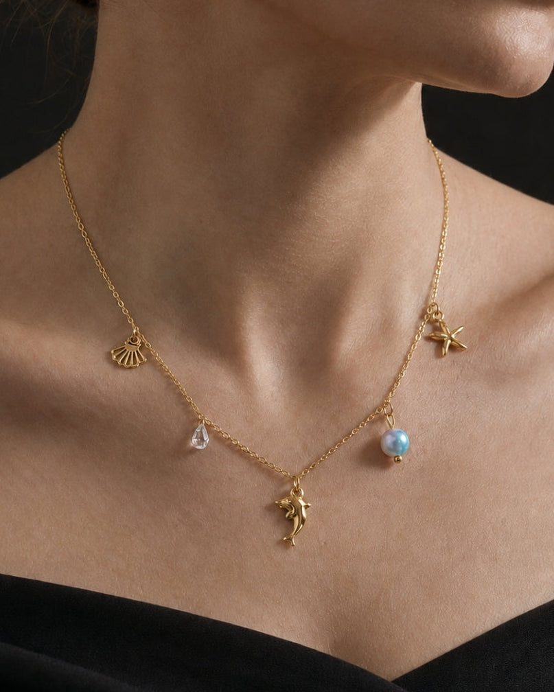 A close-up of a model wearing a delicate gold necklace with small charms, including a pearl drop and a teardrop charm, against a dark background.