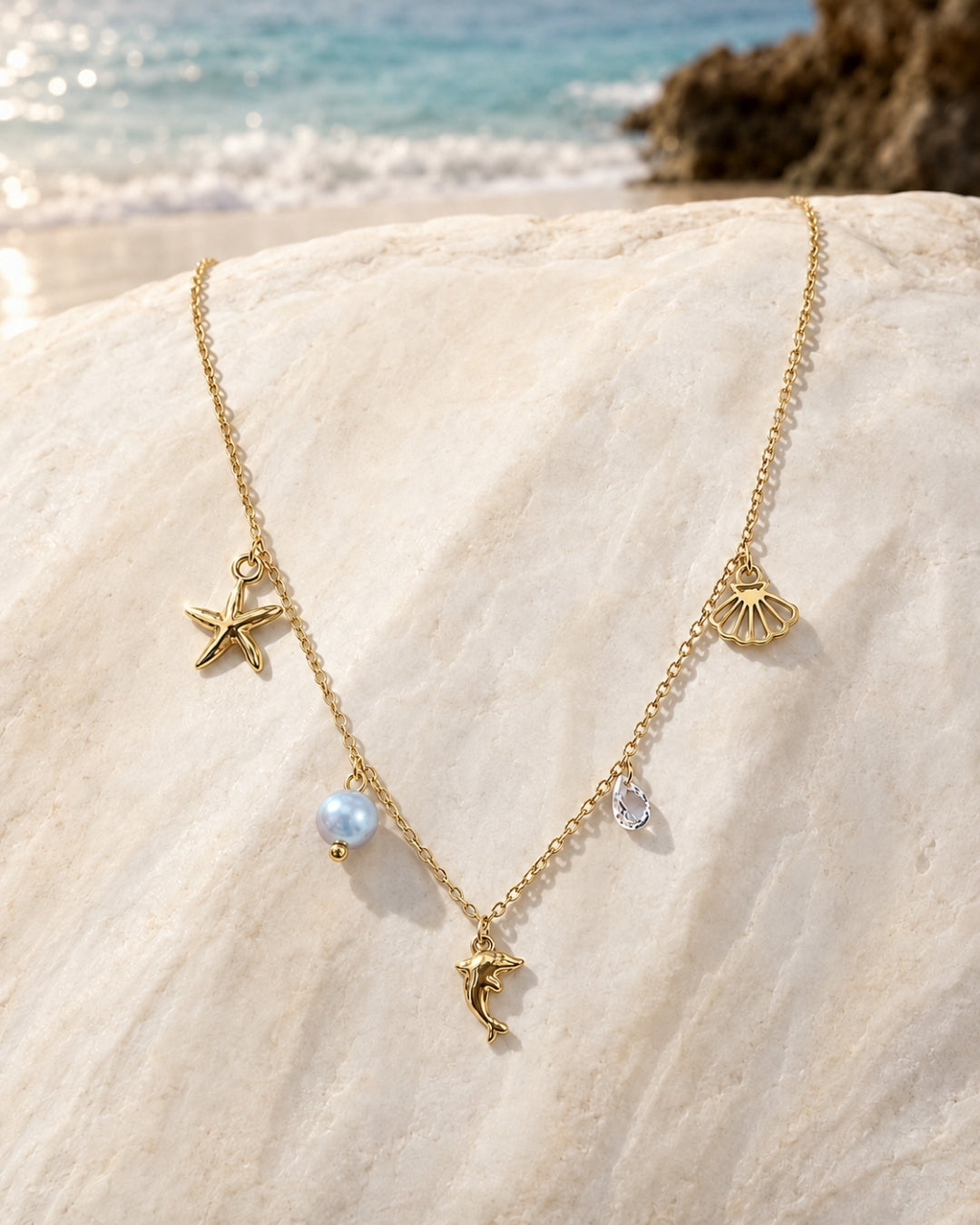 A gold-toned necklace with sea-themed charms, including a starfish, seashell, dolphin, pearl, and clear teardrop stone, draped over a large beige rock with a beach and ocean in the background.