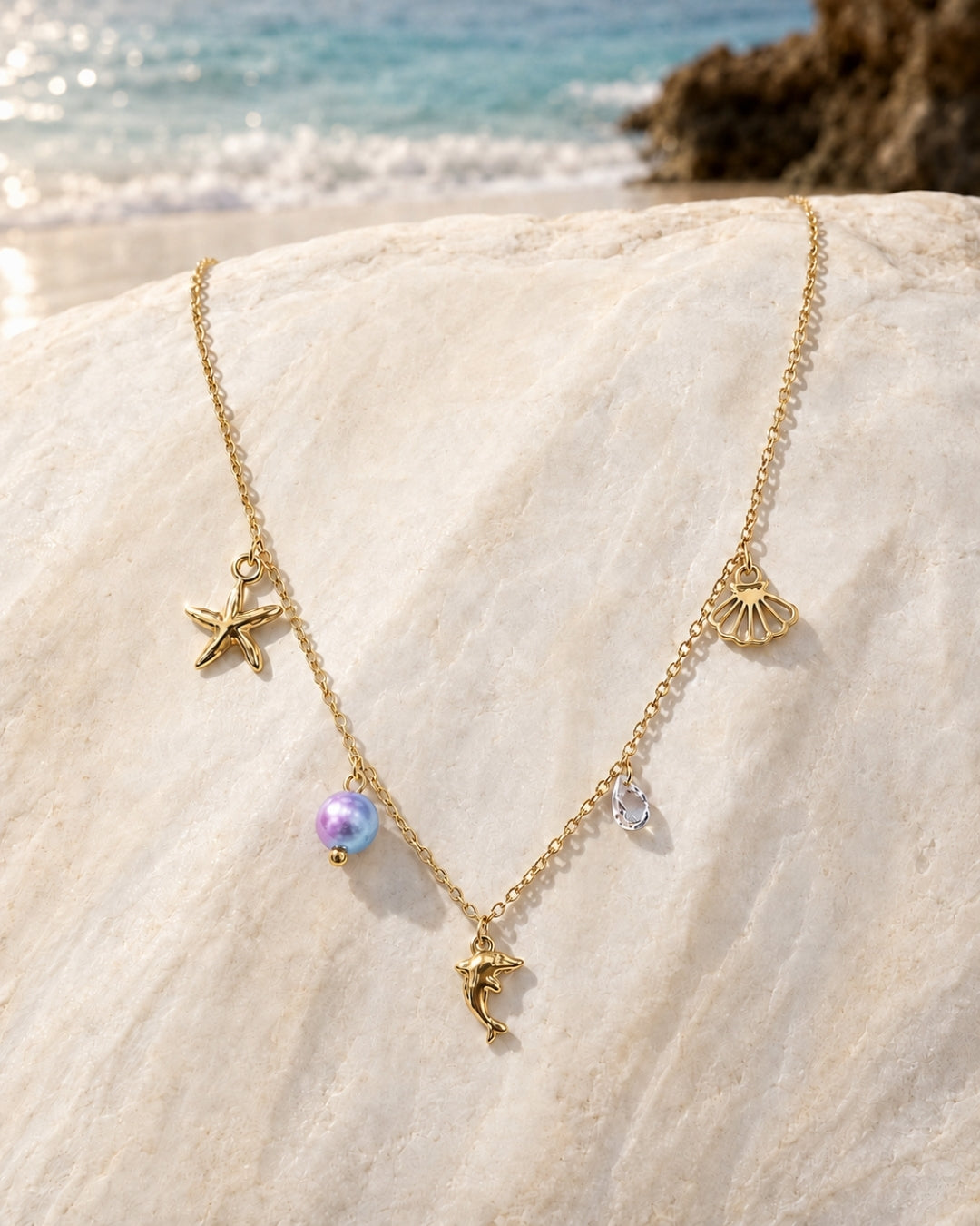 A gold-toned necklace with sea-themed charms, including a starfish, seashell, dolphin, and pearl bead, displayed on a large beige rock with a beach and ocean in the background.