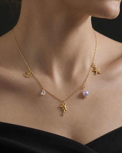 A close-up of a model wearing a delicate gold necklace with small charms, including a pearl bead and a teardrop-shaped stone, against a dark background.