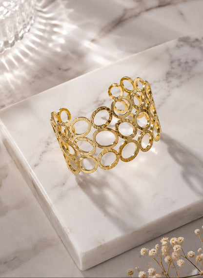 A gold-toned open-link cuff bracelet with a textured finish, displayed on a white marble block over a marble surface with soft shadows and dried flowers.