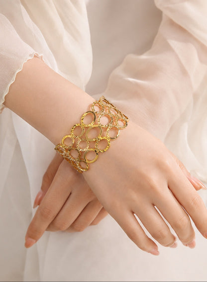 A gold-toned openwork circular link bracelet worn on a woman's wrist, shown against a soft cream-colored sheer dress fabric.