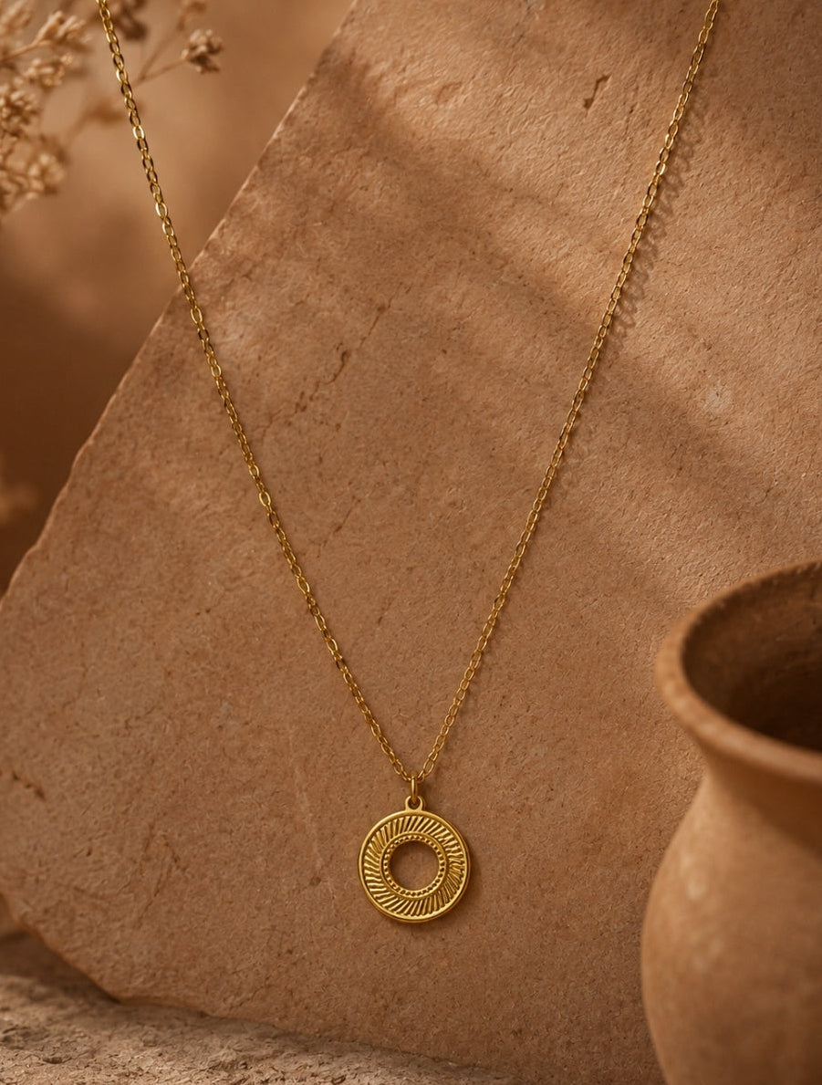 A gold necklace with a circular pendant featuring concentric textured rings, displayed on a rustic tan stone slab with dried flowers and a terracotta pot in the background.