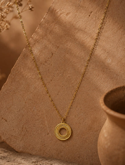 A gold necklace with a circular pendant featuring concentric textured rings, displayed on a rustic tan stone slab with dried flowers and a terracotta pot in the background.