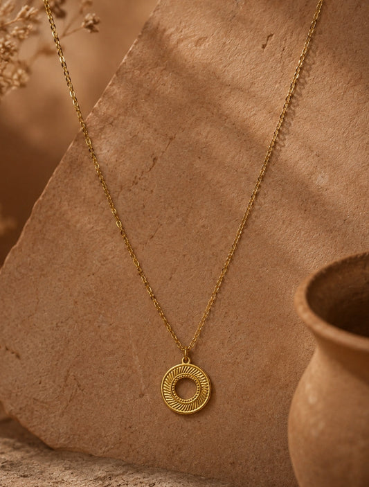 A gold necklace with a circular pendant featuring concentric textured rings, displayed on a rustic tan stone slab with dried flowers and a terracotta pot in the background.
