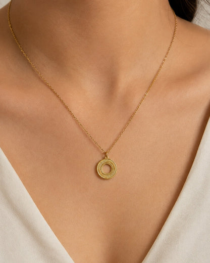 A close-up of a model wearing a delicate gold chain necklace with a textured circular ring pendant, styled with a cream-colored V-neck top.