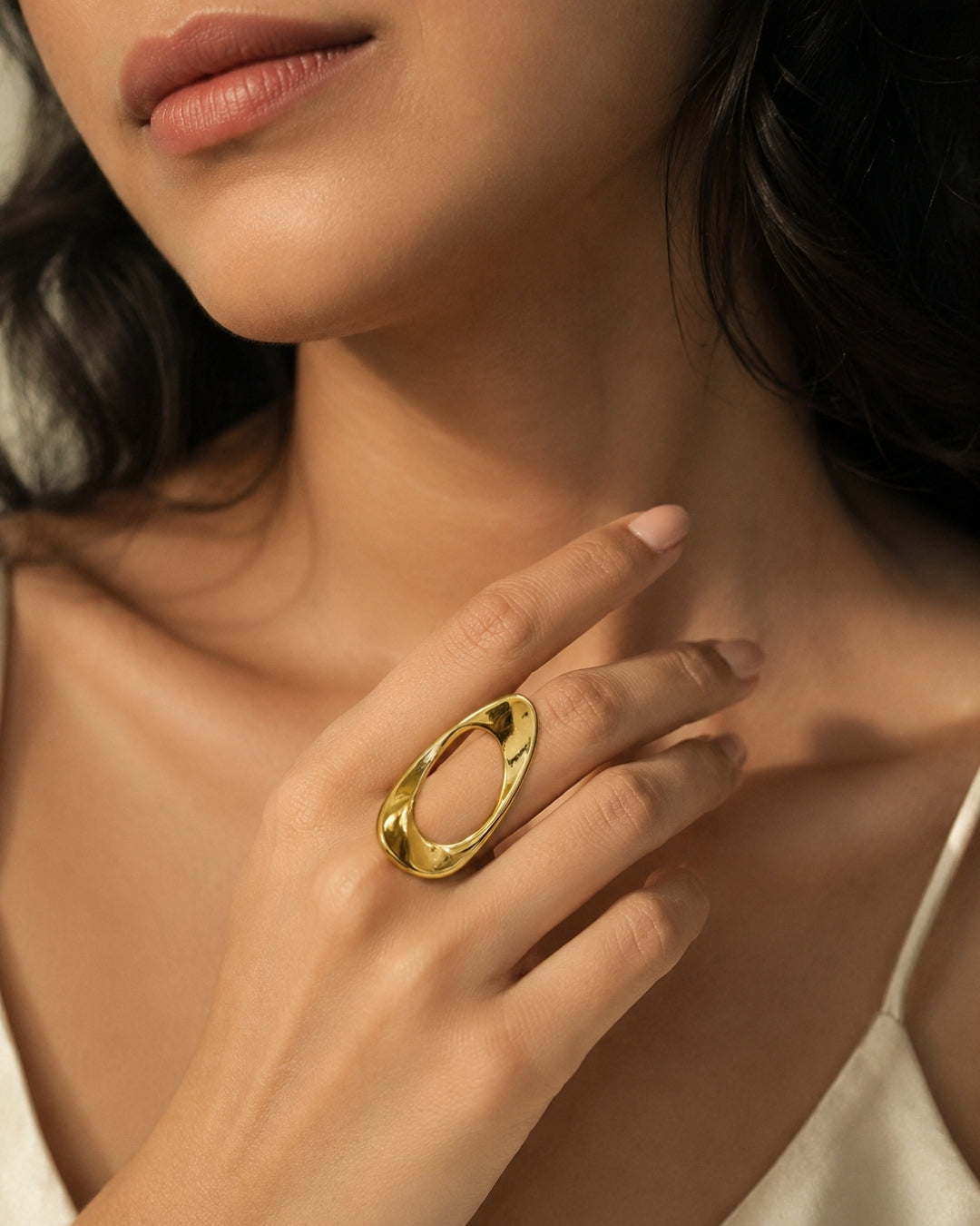 A close-up lifestyle shot of a model wearing a wide, polished gold ring with an organic, wavy open oval design on her finger.