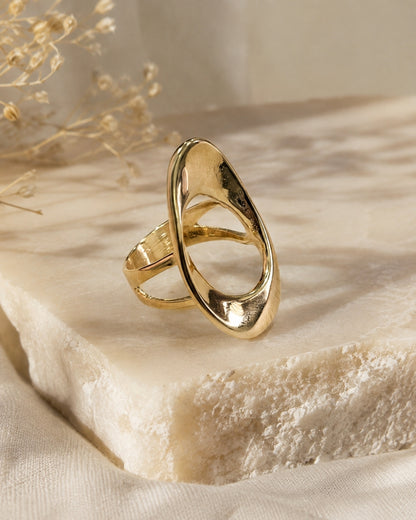 A polished gold abstract ring with a teardrop-shaped open design, resting on a textured cream-colored stone slab with dried floral elements in the background.