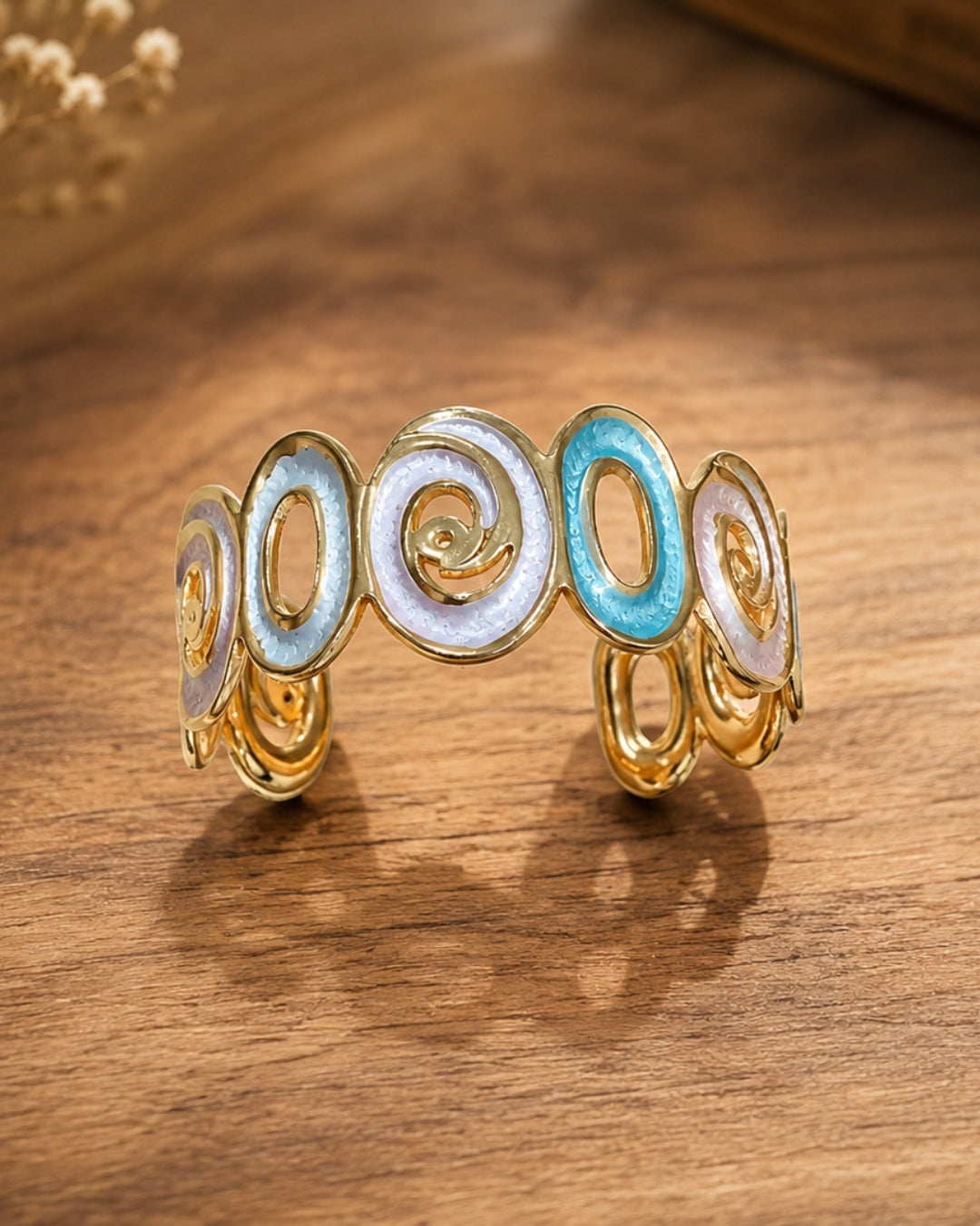 A gold-toned cuff bracelet featuring five oval links with iridescent white and light blue enamel fills, displayed on a warm wooden surface.