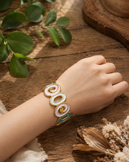 A gold-toned cuff bracelet with three large oval links filled with white enamel and small teal accents, worn on a person's wrist against a rustic wooden background with dried flowers and green leaves.