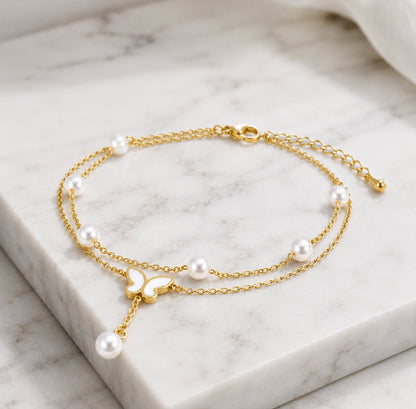 A delicate gold chain anklet with white freshwater pearls and a small butterfly charm, displayed on a white marble slab.