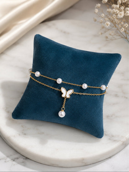 A double-strand gold chain bracelet with white pearl accents and a butterfly charm, displayed on a dark teal velvet pillow over a white marble surface.