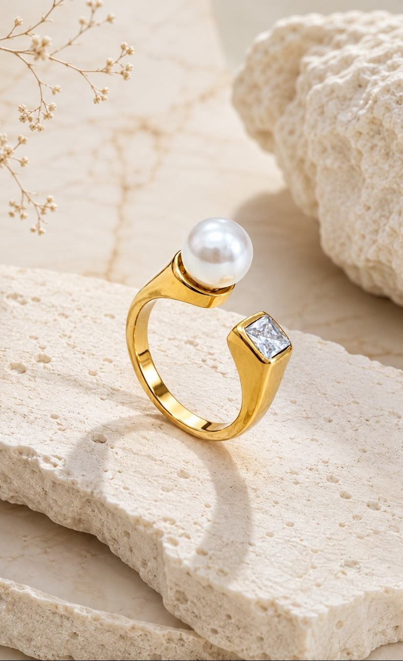 A gold ring with a round white pearl and a square clear gemstone, displayed on a textured beige stone slab.