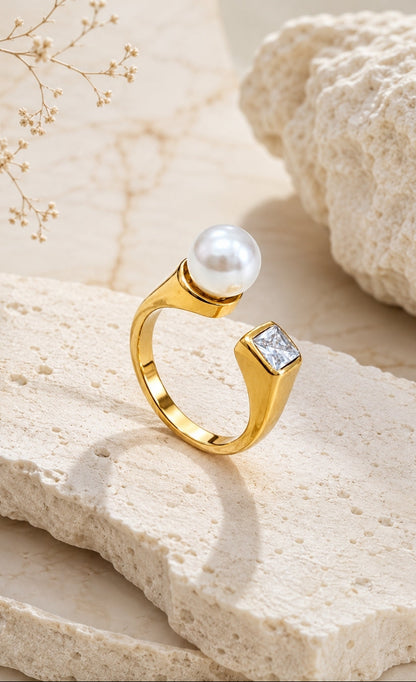 A gold ring with a round white pearl and a square clear gemstone, displayed on a textured beige stone slab.
