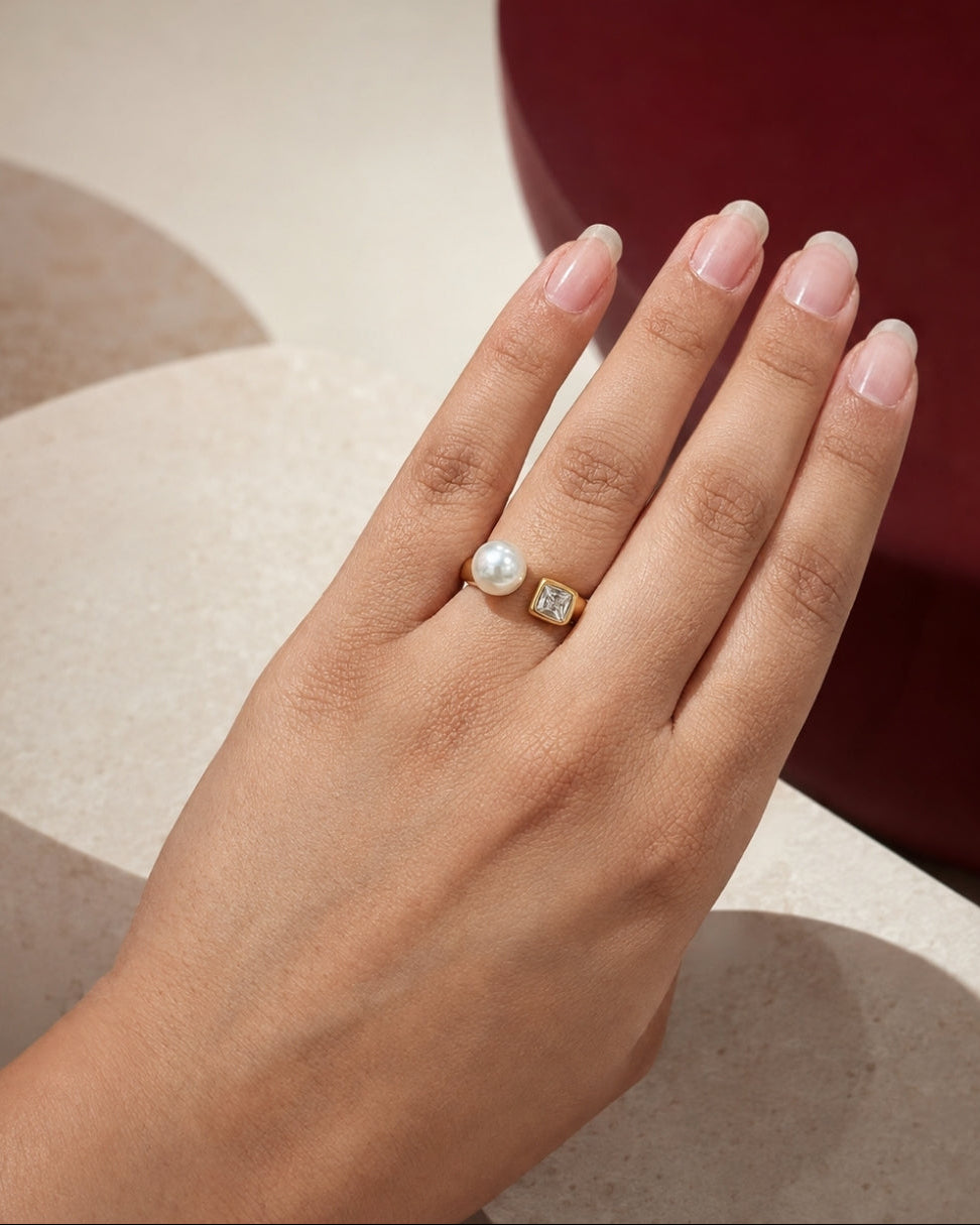 A close-up of a hand wearing two rings: a gold ring with a white pearl and a gold ring with a square clear gemstone, set against a modern background of beige geometric shapes and a dark red circular form.