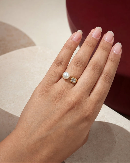 A close-up of a hand wearing two rings: a gold ring with a white pearl and a gold ring with a square clear gemstone, set against a modern background of beige geometric shapes and a dark red circular form.