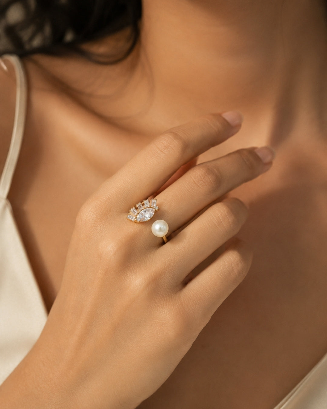 A close-up lifestyle shot of a woman's hand wearing a delicate gold ring with a single white pearl and a small cluster of clear stones above it.