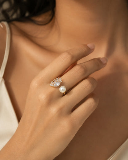 A close-up lifestyle shot of a woman's hand wearing a delicate gold ring with a single white pearl and a small cluster of clear stones above it.