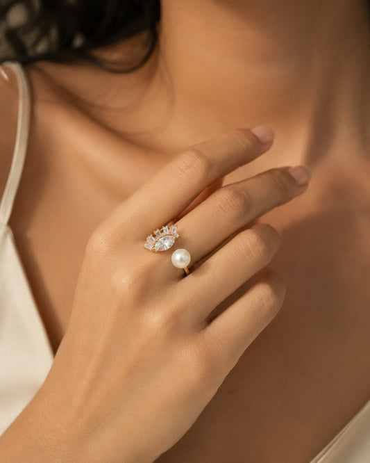 A close-up lifestyle shot of a woman's hand wearing a delicate gold ring with a single white pearl and a small cluster of clear stones above it.