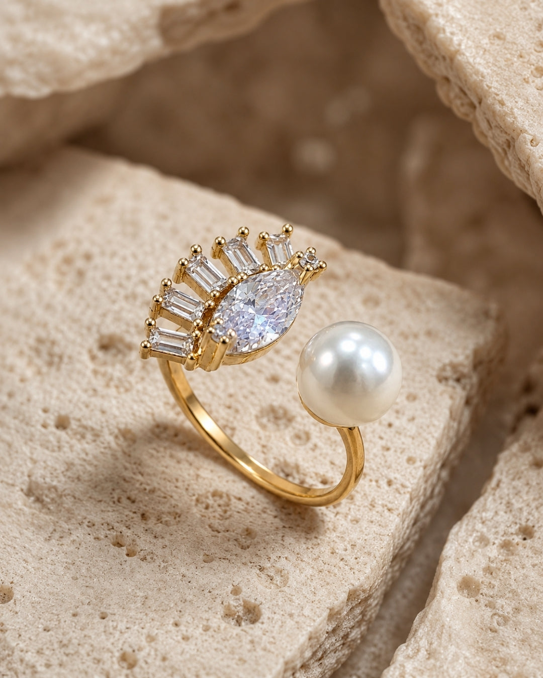 A gold ring featuring a marquise-cut clear center stone surrounded by baguette and round clear stones, with a separate white pearl, displayed on textured beige stone blocks.