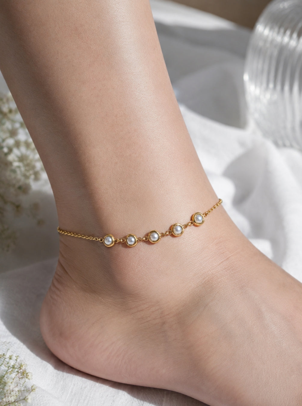 A delicate gold chain anklet with five small white pearl beads, shown worn on a person's ankle against a soft white fabric background.