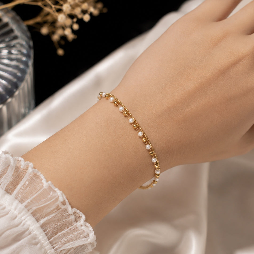 A delicate gold chain bracelet adorned with small white pearls, shown worn on a person's wrist against a soft white satin fabric background.