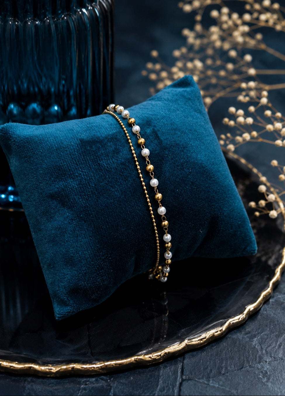 Delicate beaded bracelet displayed on a teal velvet pillow, set against a dark textured background with decorative gold-toned branches and a dark glass vessel.