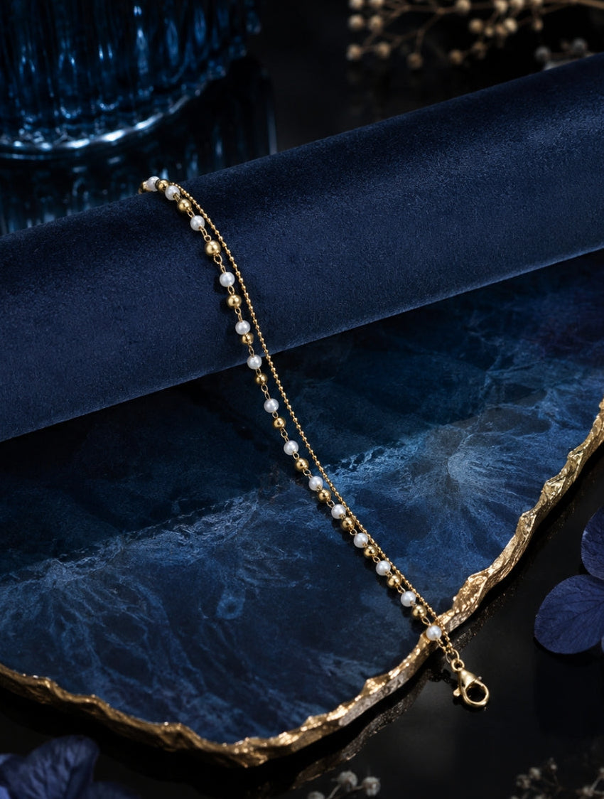 A delicate gold chain bracelet with small white pearl-like beads, draped elegantly on a dark blue velvet fabric and a gold-edged blue stone tray, styled with dried flowers.