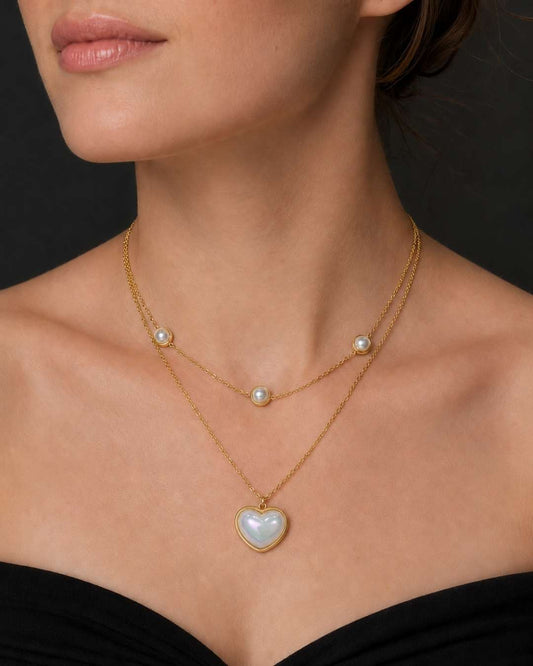 A close-up of a model wearing a delicate gold double-strand necklace with white pearl accents and a heart-shaped pearl pendant.