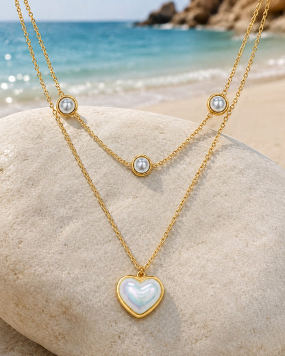 Two layered gold necklaces displayed on a large beach stone, with a turquoise ocean and rocky shoreline in the background. One necklace has a heart-shaped iridescent white pendant, and the other has three small round white pearl-like beads spaced along the chain.
