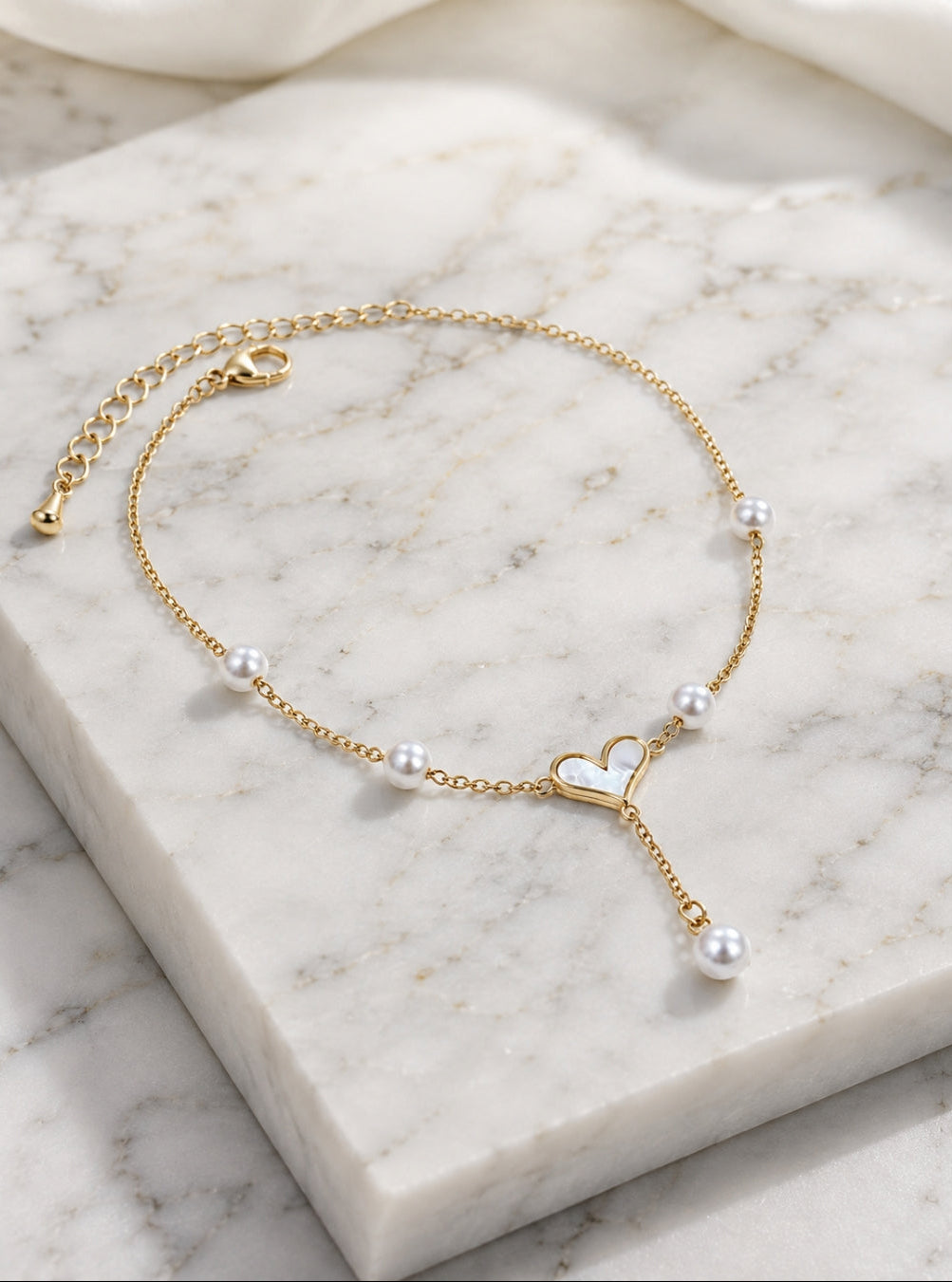 A delicate gold chain anklet with small white pearls and a central open heart charm, displayed on a white marble block.