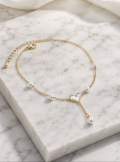 A delicate gold chain anklet with small white pearls and a central open heart charm, displayed on a white marble block.
